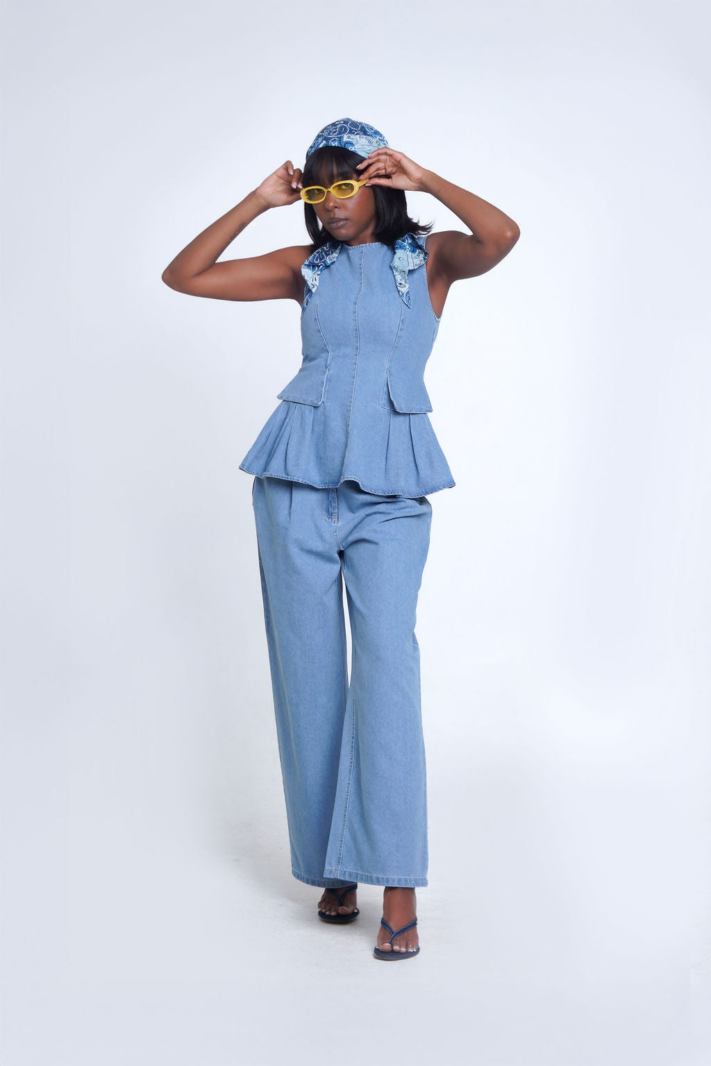 Indigo Peplum Pant Set in light wash denim — sleeveless structured peplum top with pleated ruffle hem, matching wide leg denim pants, styled with headscarf and yellow sunglasses